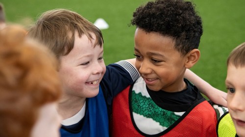Close up of a group of four young people on a sports field. They are smiling and each wearing a differently coloured sports vest