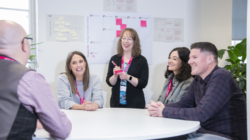 A group of five people smiling and having a meeting around a round table in an open plan office. One of them is standing up and taking notes on a Post It note