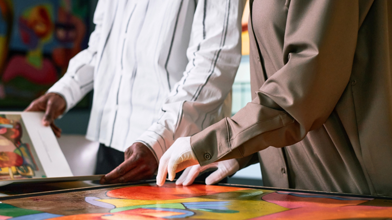 A close up of two people, one browsing through a book, the other in white gloves, hands resting on a painting 