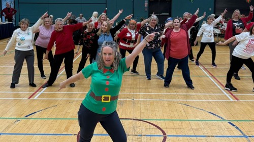  Approx 50 people taking part in a Christmas-themed dance class in a sports hall. The lead dancer is dressed as a Christmas elf and many of the participants are wearing Christmas t-shirts and accessories