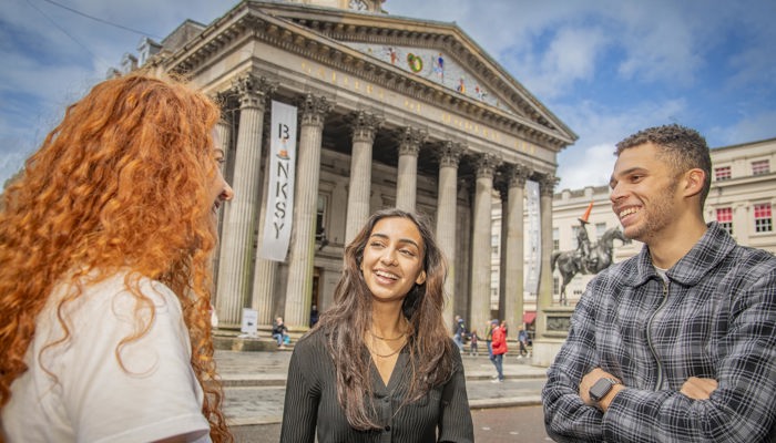 Three people smiling while standing in front of the Gallery of Modern Art in Glasgow on a sunny day.