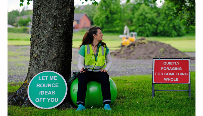 The artist sits on a green yoga ball with a high vis vest. To there left a green sign reads "let me bounce ideas off you".