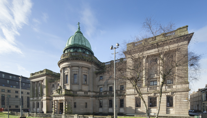 The Mitchell Library, Glasgow, on a sunny day. A large light sandstone building, with many windows and statues on the architecture of the building. Grass and trees are in front of the building.