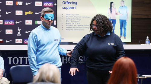 A person is leading another person who is blindfolded. They have their arm outstretched and the blindfolded person is holding the back of their arm. There are both stood in front of a group of people at a training session