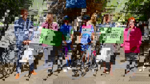 A group of six people gathered for a photo in front of large archway in a city park. Two people are wearing cycling clothes and helmets and are standing next to racing bikes. Two are holding signs that read Lloyds Tour of Britain