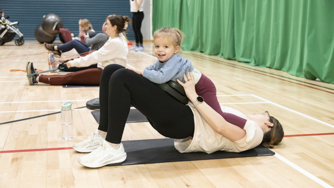 A parent performs a glute bridge exercise on a mat while a smiling toddler sits on their hips, during a parent-and-child fitness class in a sports hall.