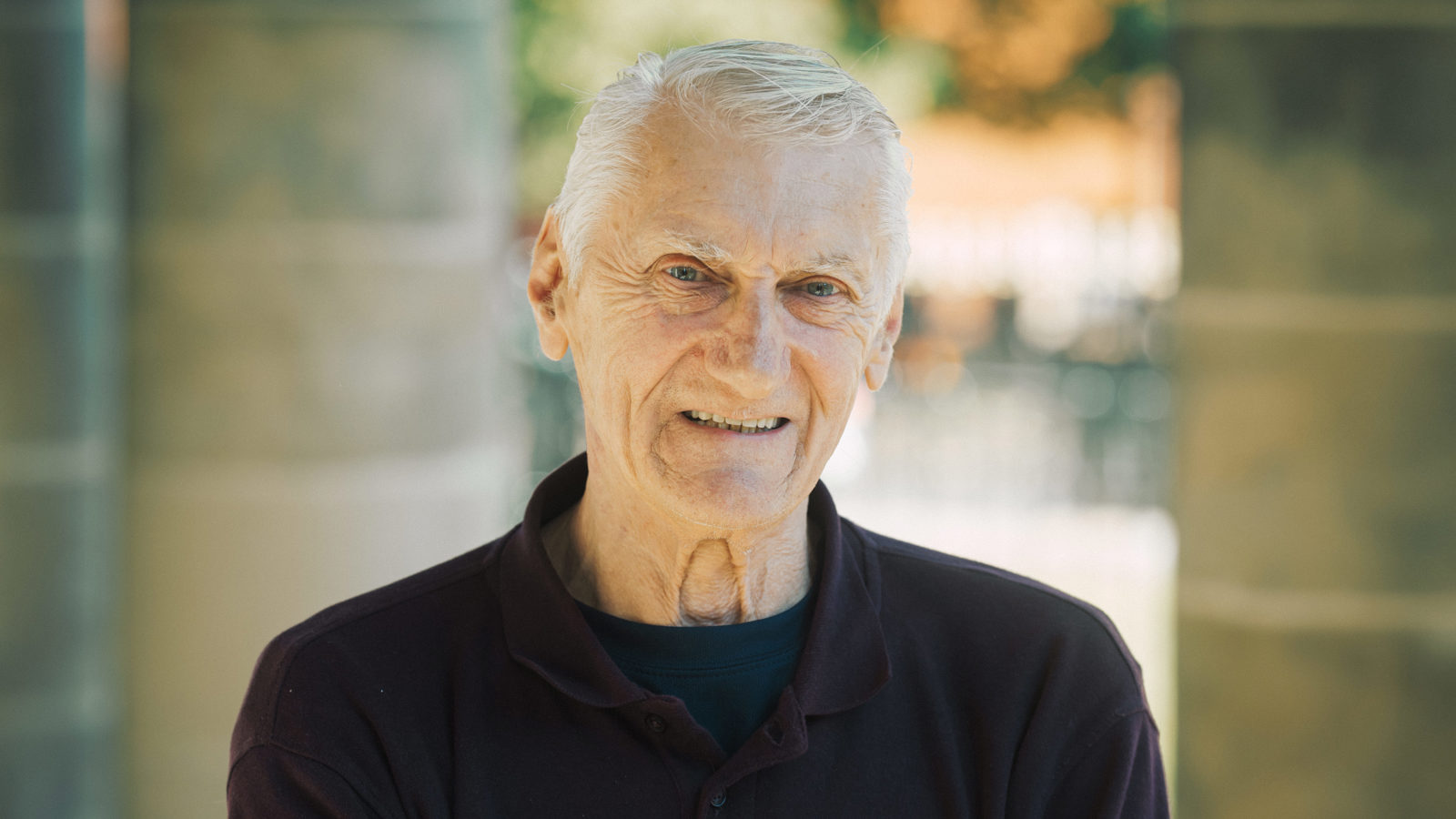 A man with grey hair stands smiling with his arms folded in front of a pillar