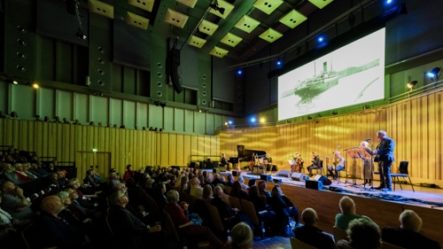 An audience watching performers on a stage. There are two people standing at microphones, a celloist and pianist. Behind them is a screen showing an black and white image of a steamer ship
