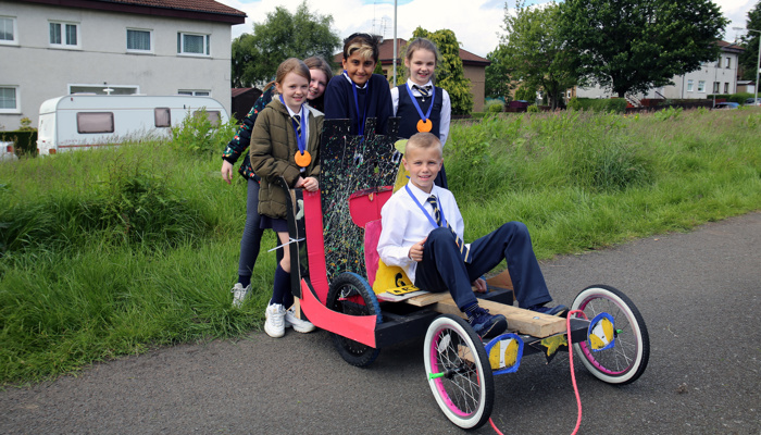Children show off their hand made racing cart. The cart is a bright red and has three wheels.
