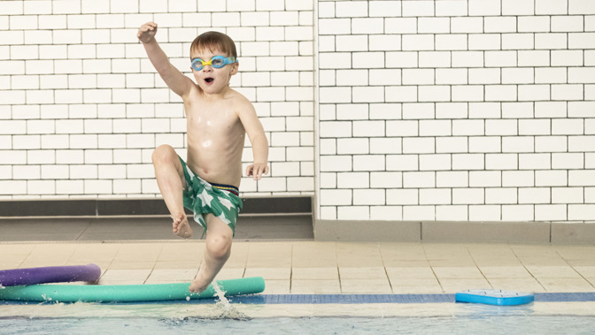 A little boy wearing swimming trunks and armbands gives a triumphant punch to the air as he leaps into a swimming pool