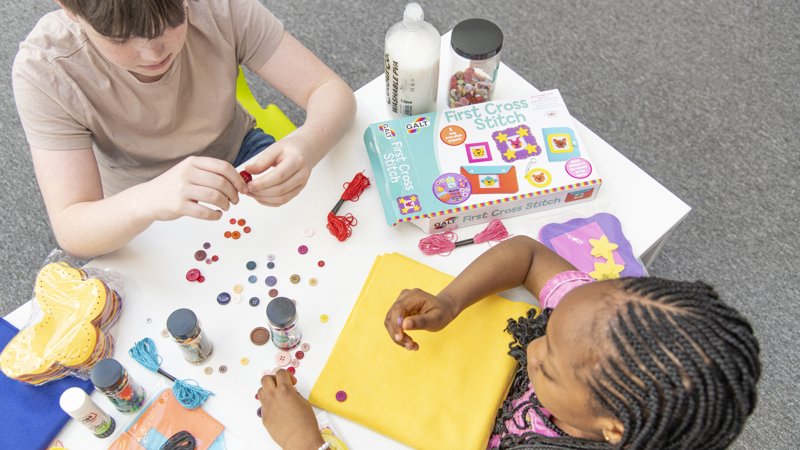 Looking down from above over two children sitting at a crafting table, which has colourful buttons, paper, string and more.