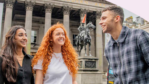 Three people smiling, standing outside a city centre museum. Behind them is a statue of a person on a horse. The statue has a traffic cone on the person's head.