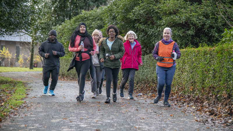 A diverse group of people jogging on a path in a park with bushes on one side of them.