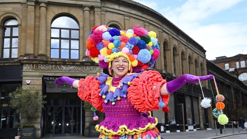 A person wearing a colourful knitted costume and a large pom pom hat, smiling with arms outstretched outside a large stone city building