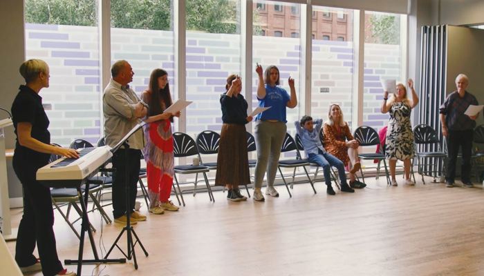 A group of people standing up during a singing for fun session, which is being led by an instructor who is standing in the corner of the room at a piano.