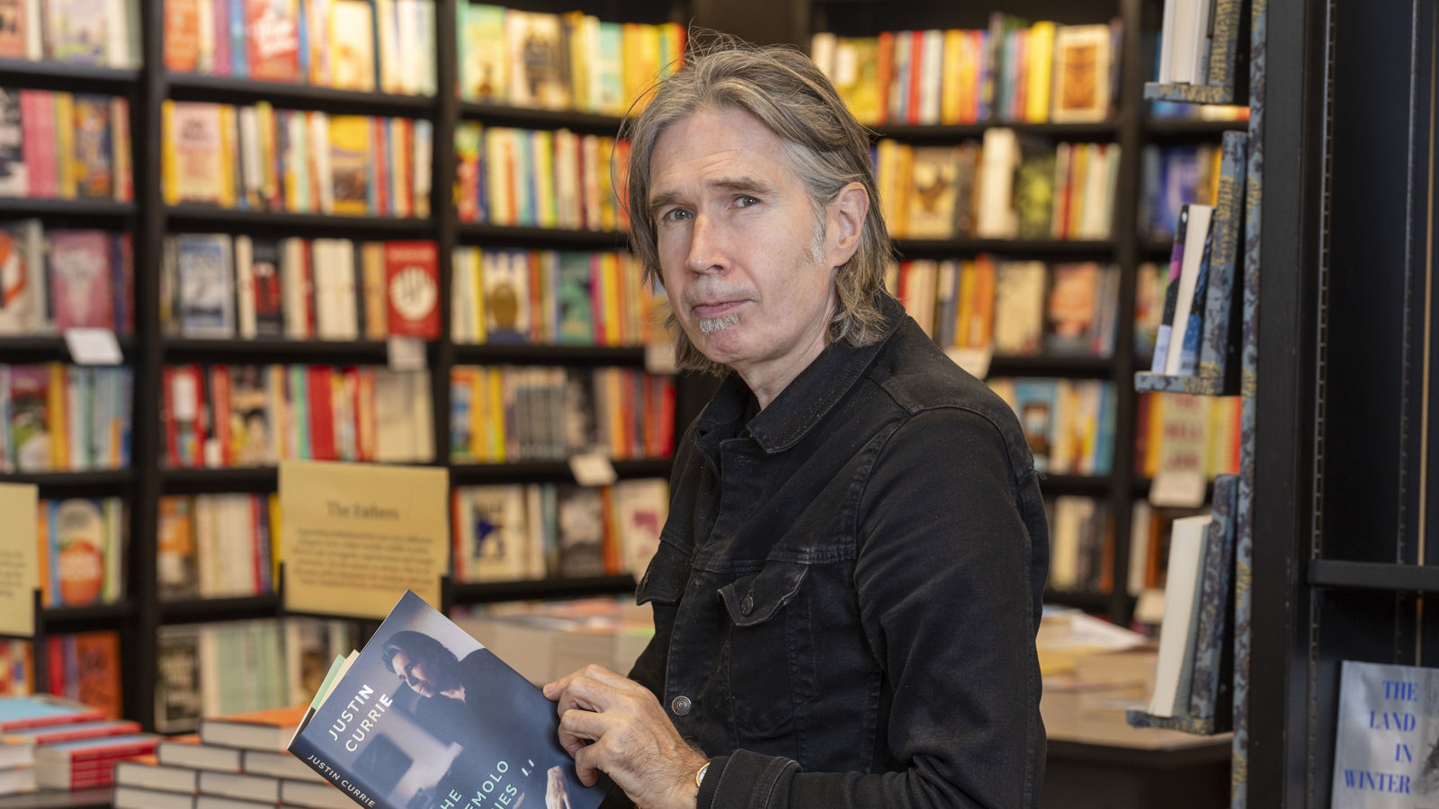 A man with long dark hair opens a book in a well-lit library