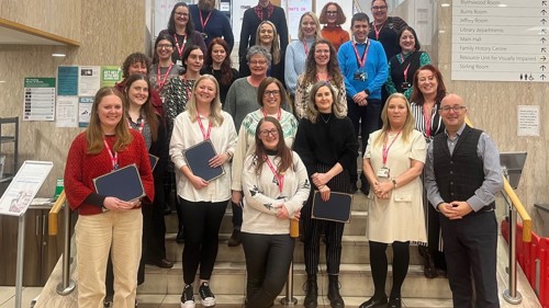 A large group of people gathered for a graduation photo on a wide staircase in a library. Some of them are holding certificates