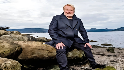 A person in a dark double breasted coat, sitting on a large rock on a loch shore