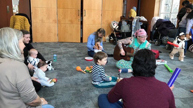 Children and parents are gathered around a person sitting on the floor playing a guitar