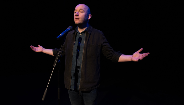 A performer wearing dark clothing stands in front of a microphone with their arms outstretched