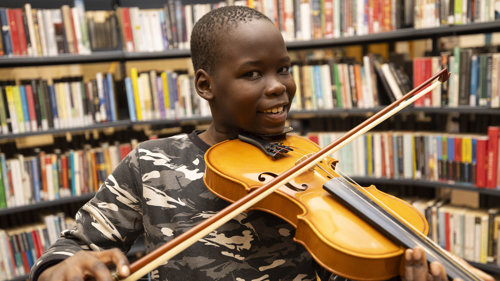 A young boy smiling while holding a violin and bow in front of shelves full of books inside The Mitchell Library in Glasgow.