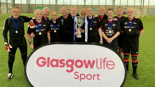 A football team made up of older people all wearing the same black kit and posing for a photo with a large silver in an indoor football centre