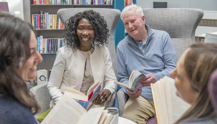 A smiling black lady and smiling white man sit in beauitful grey chairs in a library, with books in their hand, enjoying a conversation
