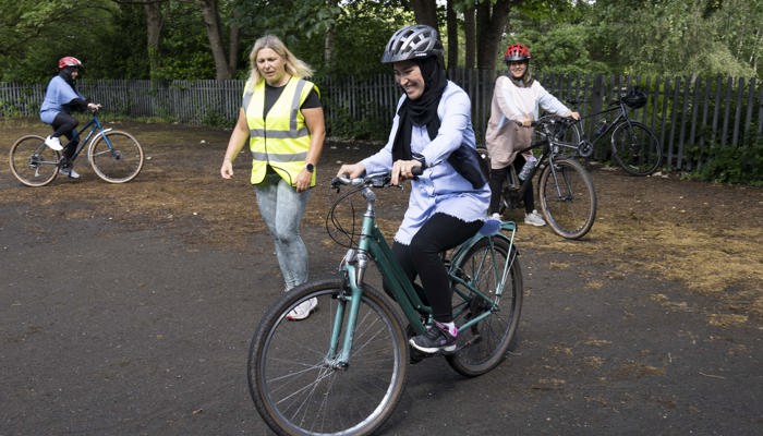 A young lady with a big smile on her face learning to ride a two wheeled bicycle with help from a coach wearing a high vis yellow vest