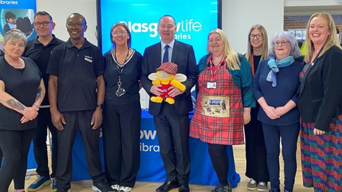 A group of nine people are gathered for a photo in a community centre. They are standing in front of a TV screen displaying text Glasgow Life Libraries
