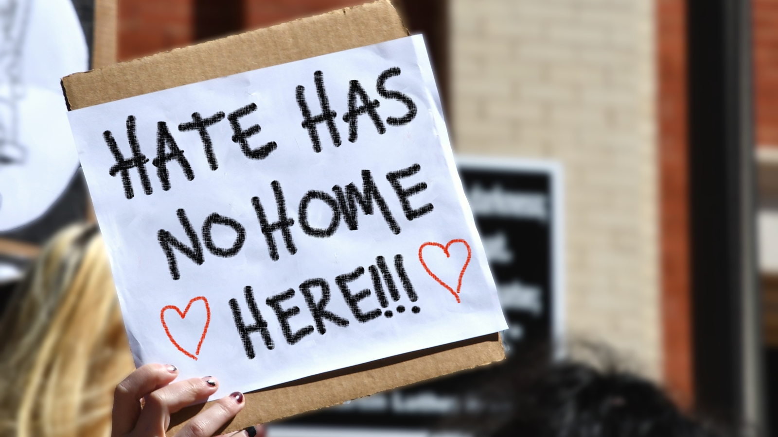 A person holding a protest sign that reads “Hate Has No Home Here!!!” with two red heart drawings, in an outdoor urban setting.