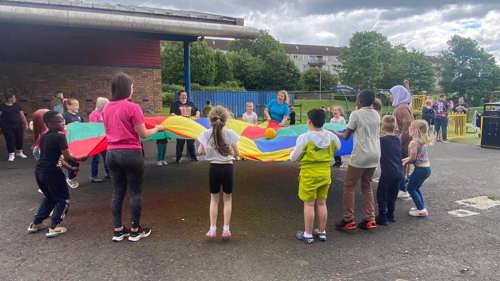 A group of adults and young people gathered around a large colourful circle of material in a playground. They are stood in a circle and everyone is holding the material in their hands