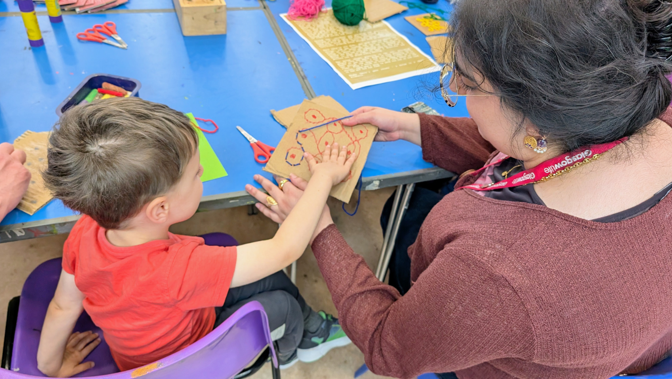 A young child interacts with an adult about crafts as they sit at a colourful table