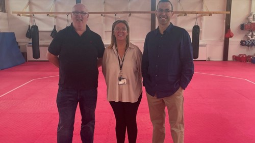 Three people standing and smiling in a gym area of a community centre. There are punching bags on the wall behind them.