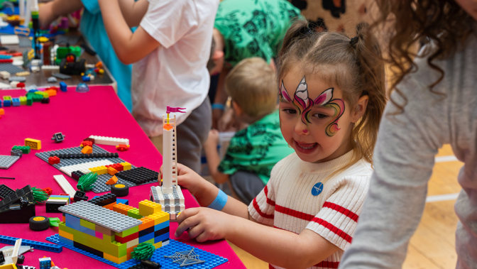 Children with painted faces play with LEGO bricks on a table covered in colorful pieces at an indoor activity event, with adults supervising in the background.
