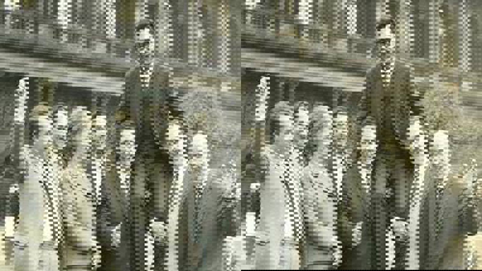 One person wearing glasses smiling while sitting on the shoulders of two people, with another 5 people celebrating with them by waving hands in the air and smiling. There is a large building and shrubs in the background.