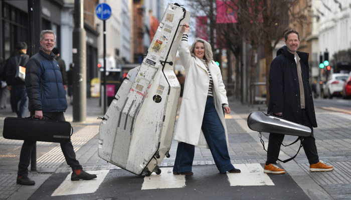 Three people walk across a street carrying musical instruments
