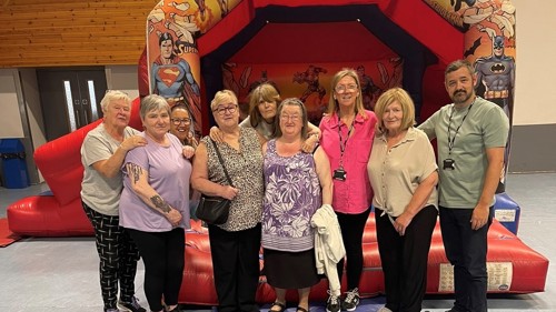 A group of nine people standing in front of a bouncy castle in a community centre hall. They are smiling and some have their arms around each others shoulders