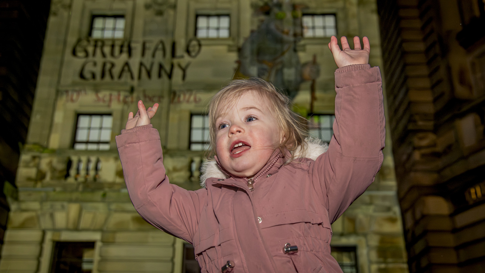 A young person wearing a pink coat is outside a large  Edwardian library building in the evening. There arms are outstretched and there is a light projection of the Gruffalo character behind them
