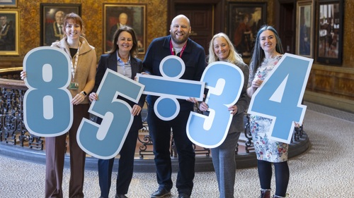 A group of five people posing for a group photo in a marble hallway. They are holding large cutout numbers and mathematical symbols