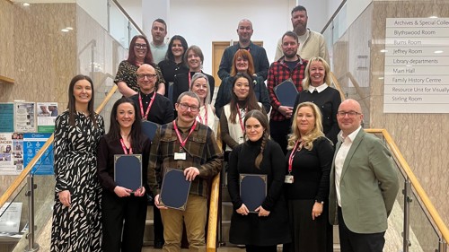 A group of 18 people gathered for a photo on a marble staircase in a library. Some of them are holding certificates