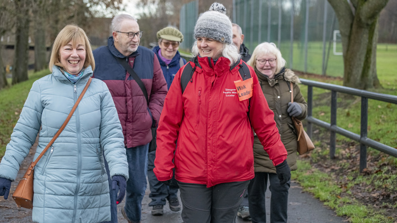 A smiling group of senior citizens on a health walk, led by a woman in a red waterproof jacket with a 'Walk Leader' badge.