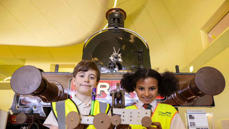  Two schoolchildren wearing yellow safety vests pose in front of a historic steam locomotive inside a museum, smiling as part of an educational visit