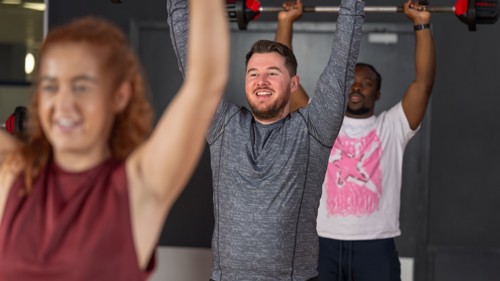 Three Glasgow Club members taking part in Body Pump class they all are smiling and are holding weighted barbells over their heads.
