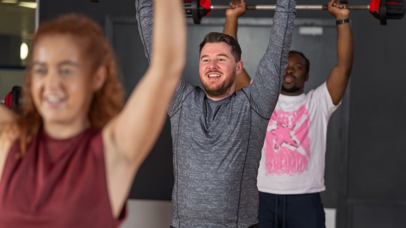 Three Glasgow Club members taking part in Body Pump class they all are smiling and are holding weighted barbells over their heads.