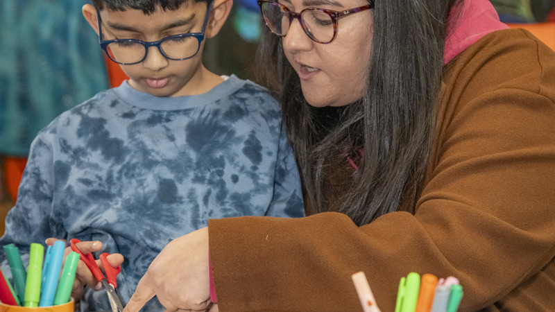 A young boy and his mum in a multi-coloured room during an art club  using red scissors to cut paper, they are sitting at a blue table with pens and glue sticks on it