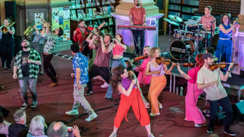 A large group of musicians and singers in colourful clothes performing energetically to an audience in a library