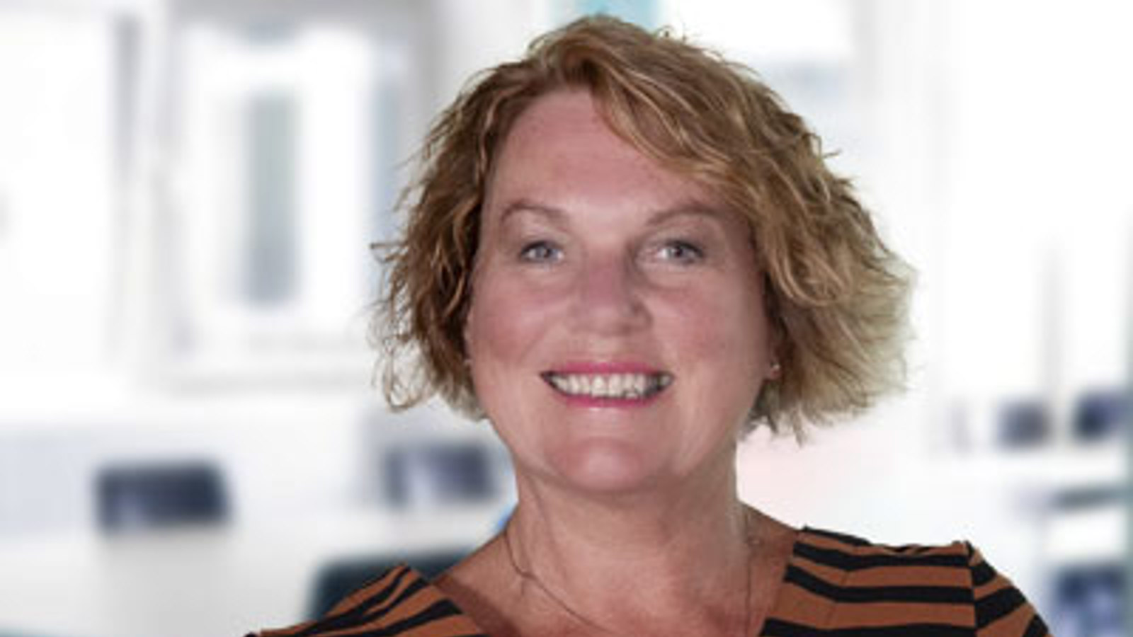 A headshot of Jan Buchanan, a person with blonde wavy hair wearing a brown and black striped top