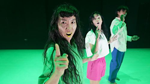 Three people standing in a line taking part in a dance performance in a green space. They are wearing loose colourful shirts, trousers and shorts and have one hand raised with index finger pointed