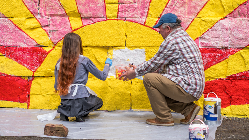 A child and adult paint a mural on a wall. It shows a setting sun, it is bright yellow and pink.