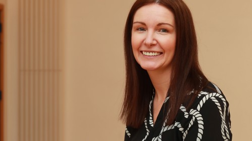 A woman with dark, shoulder-length hair wearing a black and white top is smiling as she faces the camera side-on